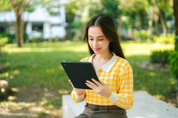 Obraz premium A woman in yellow checkered sweater sits outdoors, using tablet in park. Digital Nomad Lifestyle Freelance Hybrid Work communicating with a colleague