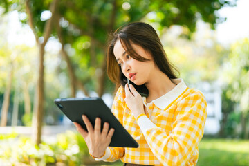 Fototapeta premium A woman in yellow checkered shirt is using tablet while talking on phone in sunny park. Digital Nomad Lifestyle Freelance Hybrid Work communicating with a colleague
