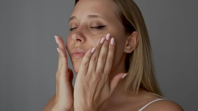 Young blonde woman massaging her face quickly tapping herself on the cheeks to improve blood circulation, relax the muscles, tighten skin on dark grey background. Cosmetology and beauty concept