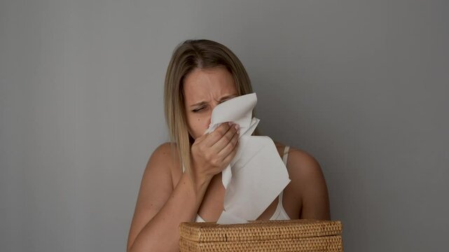 A young caucasian woman sneezing from allergy covering the nose with a facial tissue and then throwing it aside on a dark grey background. Disease, virus, flu, cold, seasonal allergy, runny nose