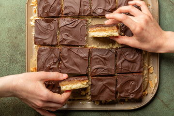 Girls hand taking a piece of Caramel shortbread, also known as caramel squares, slice, or millionaire's shortbread. Dessert, cake made with  shortbread biscuit base topped with caramel and chocolate.