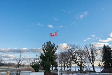 Canadian flag waving proudly against a vibrant blue sky, overlooking a tranquil waterfront scene.  Bare winter trees frame the picturesque view, hinting at the season's peaceful beauty.