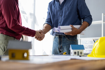 Architect holding blueprints is shaking hands with a client in his office during a meeting about a real estate project.