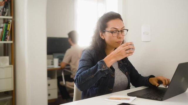 Focused Latin woman multitasking with laptop, taking notes, and sipping coffee while working from home. Partner in the background also engaged in remote work.