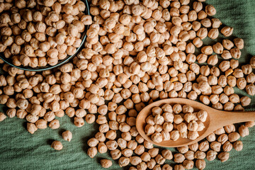 Top view of scattered dried chickpeas on green fabric with a wooden spoon and a ceramic bowl. Rustic setup highlighting healthy eating, plant-based diets, and natural food ingredients