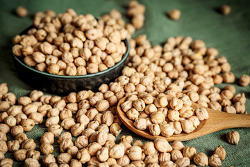 Rustic close-up of dried chickpeas in a wooden spoon and a bowl on a green fabric background. Natural light highlights textures, perfect for healthy food, vegan recipes, and organic lifestyle concepts