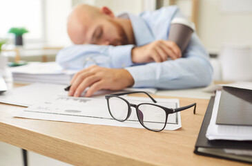 Fototapeta premium Tired sleeping office man, employee falling asleep at desk overworked, exhausted sleeps stressed male manager lying on table and sleeping after hard working day, no energy left, glasses put away