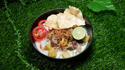 Soup or Soto Betawi with savory coconut milk broth filled with pieces of potato and beef is a traditional Indonesian food typical of Jakarta. Served in a black bowl on synthetic green grass
