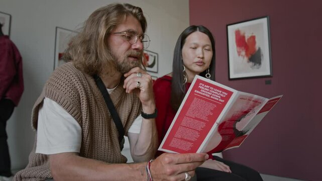 Low angle shot of bearded Caucasian man and Asian woman sitting closely together in art gallery, reading leaflet, while another person looking at abstract artwork on walls behind them