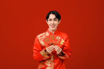 A smiling young Asian man in a red and gold traditional Chinese outfit holds festive red envelopes,...
