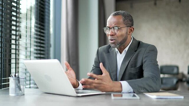 Upset worried african american businessman reads bad news on laptop sitting at desk at workplace in business office. Frustrated entrepreneur is sad while reviewing negative information on a computer