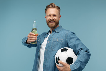 Positive bearded blonde hipster man holding a bottle of beer and classic soccer ball over blue background in studio