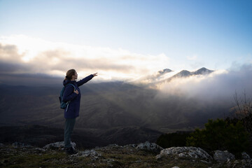 A side view of a hiker woman looks at the view with a raised hand on top of the mountain at the sunset sunrise. A girl with a backpack stands on a cliff's edge and points the way. Active lifestyle.