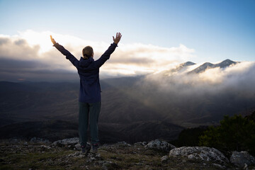 An unrecognizable traveler woman with raised hands on the cliff's edge in the sunset. Hiker girl with hands up on top of the mountain wear hiking clothes. Win, achieving outdoors. Backside view.