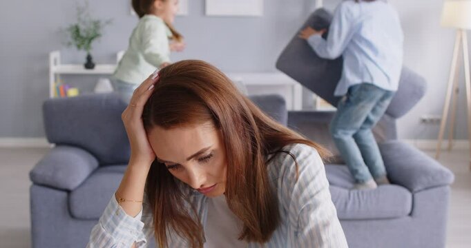 Tired upset unhappy stressed desperate mother does not know how to deal with hyperactive children who are having fun, jumping on sofa, fighting pillows and making noise. Parenting naughty kids concept