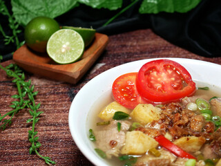 Clear beef soup served in a white bowl with a blurred background of lime, green leaves and black fabric, on a brown wood pattern background. Close up and Selective focus