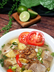 Clear beef soup served in a white bowl with a blurred background of lime, green leaves and black fabric, on a brown wood pattern background. Close up and Selective focus