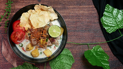 Top view Soup or Soto Betawi with coconut milk broth filled with potato slices and beef is a typical food of Jakarta Indonesia. Served in a black bowl on a brown background decorated with green leaves