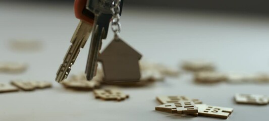 Male hand dangling keys with a keychain in the shape of a house with small wooden house models in the background - Powered by Adobe