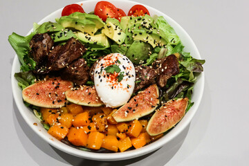 Close up of poached egg, pumpkin, beef pieces, avocado, cabbage and assorted vegetables salad with black and white sesame  in a white bowl