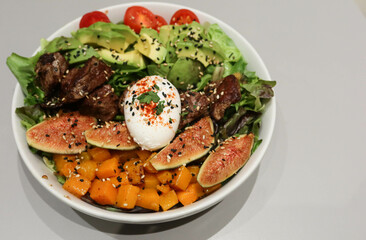 Close up of poached egg, pumpkin, beef pieces, avocado, cabbage and assorted vegetables salad with black and white sesame  in a white bowl