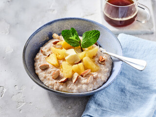 A delicious bowl of oatmeal topped with fresh pineapple chunks, sliced almonds, and mint leaves sits on a light table. Beside it, a steaming cup of herbal tea adds warmth to the morning