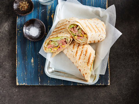 A well-prepared grilled wrap filled with lettuce, meats, and toppings rests on a rustic wooden board. Accompanied by salt and pepper, this dish looks appetizing and satisfying - Powered by Adobe