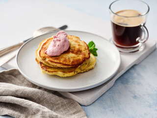 A stack of fluffy pancakes with a dollop of pink cream sits on a plate next to a cup of dark coffee. The breakfast is displayed on a light blue table with a graceful cloth