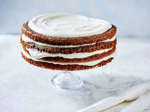 A beautifully constructed cake features multiple layers of spiced cake and generous cream frosting, displayed on a clear cake stand in a light and inviting kitchen space