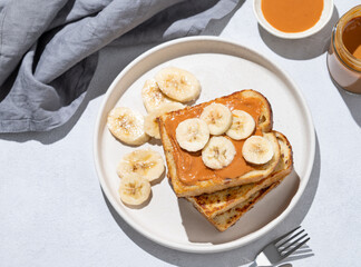 Peanut butter toast with banana on a plate on a light background with napkin and shadow.