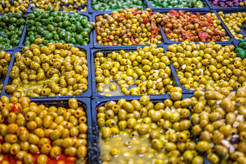 A vibrant market display of various marinated green and yellow olives in trays, seasoned and colorful