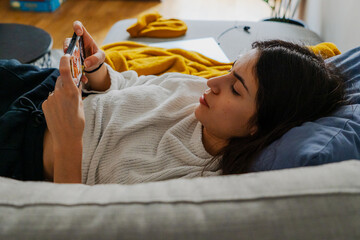 A relaxed young woman in casual attire lounging on a plush sofa, scrolling through her phone with a serene expression on her face.