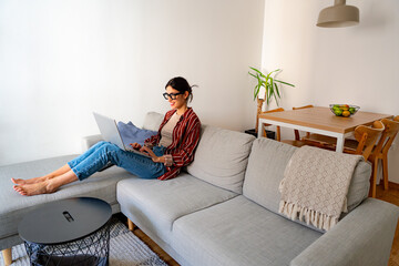 Obraz premium Smiling woman enjoying a peaceful afternoon on her couch, working on her laptop in a bright, modern space. Copy space