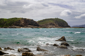 Rocks bathed by the sea with mountains in the background and partly cloudy sky. Photographed on Conchas beach, coast of Rio de Janeiro, Brazil.
