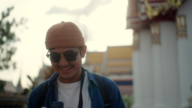 Happy young asian man using camera and enjoying culture landmark travel in temple Thailand, solo traveler capturing ancient pagoda memories on their adventure, tourist hipster camera photography.
