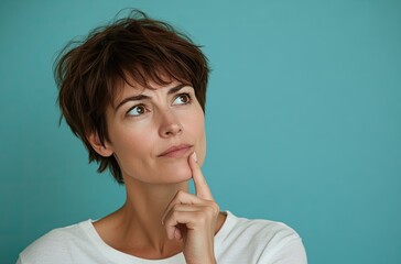 A woman in her late thirties with short brown hair, wearing a white t-shirt, is thinking while holding one finger on her chin and looking to the side over a light blue background with copy space
