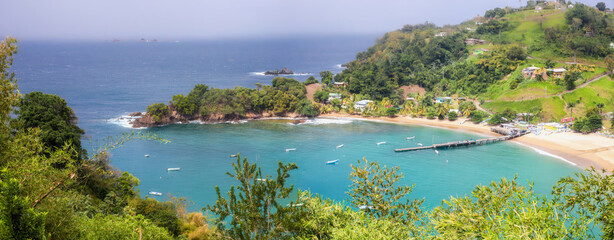 Obraz premium Panoramic view from above to Parlatuvier Bay with crystal clear water, green hills, and charming coastal homes. Wooden pier and small fishing boats . Tropical escape on Tobago, Trinidad and Tobago.