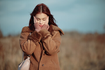 Teenage girl with red hair and glasses, hands on cheeks, looking thoughtful and slightly anxious, dressed warmly in a brown coat on a breezy autumn day.