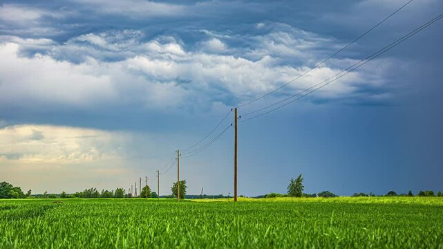 Electricty pylon tower poles on a sprawling green field in Latvia, with dramatic storm clouds looming above as cars drive on country road