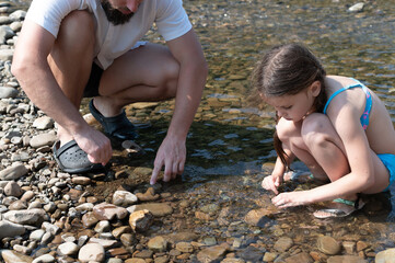 A man and young girl squat by a clear stream, carefully observing the rocks and pebbles at the...