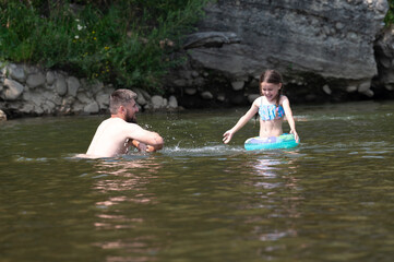 Fototapeta premium A man and young girl are playing in a calm river. The girl floats on a small inner tube. The rocky riverbank is visible in the background. It's a sunny day, and the water appears cool and inviting.