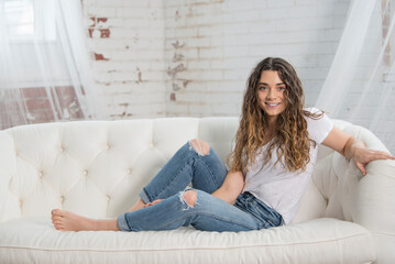 Obraz premium Beautiful young brunette woman relaxes on white couch wearing white t-shirt and blue jeans