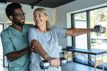 Obraz premium African American doctor helping patient treating arms with dumbbells during rehabilitation session