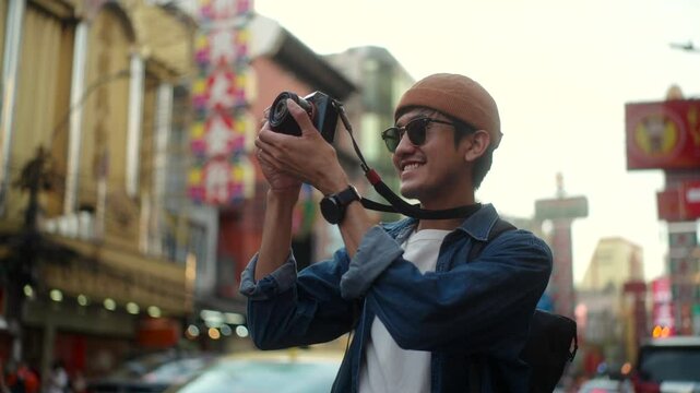 Happy young asian traveler man wearing sunglasses and hat using camera to take photo while standing in famous china town bangkok street in Thailand, solo trip lifestyle concept.
