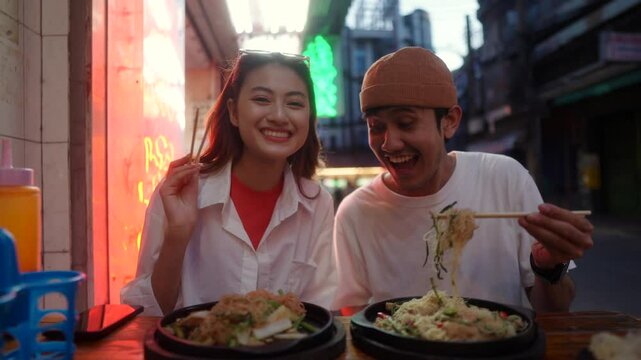 Happy young couple enjoying food and local noodles together at a street food market in Bangkok, two asian people sharing fun moments and love while exploring food culture in a restaurant.
