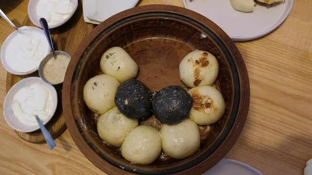 Ukrainian dumplings, or Galushki, with various fillings served in a clay pot alongside sour cream and sauce, hands taking one dumpling to plate for eating.