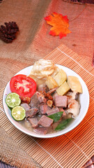 Closeup of a bowl of clear beef soup with chunks of beef, potatoes, fresh tomatoes, lime slices, and fried onions. Served on bamboo mats, burlap, and leaves.