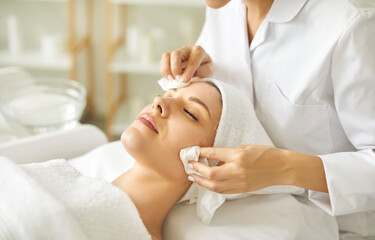 Relaxed young woman enjoying facial treatment at spa salon. Professional beautician or cosmetologist in white coat wipes off makeup or refreshing AHA peel from woman's face. Skin care, beauty concept