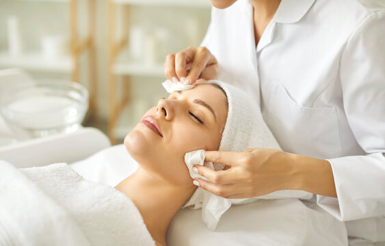 Relaxed young woman enjoying facial treatment at spa salon. Professional beautician or cosmetologist in white coat wipes off makeup or refreshing AHA peel from woman's face. Skin care, beauty concept