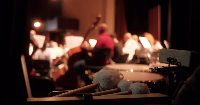 A warm, atmospheric view of percussion mallets and instruments in the foreground, with an academic classical orchestra in rehearsal visible in the blurred background